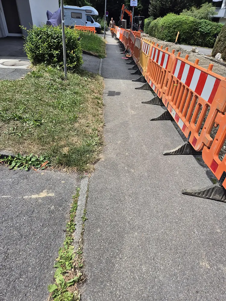 Orangefarbene Absperrungen blockieren den Gehweg in der Nähe von Grünflächen, die von der Grauflächenpflege betreut werden.